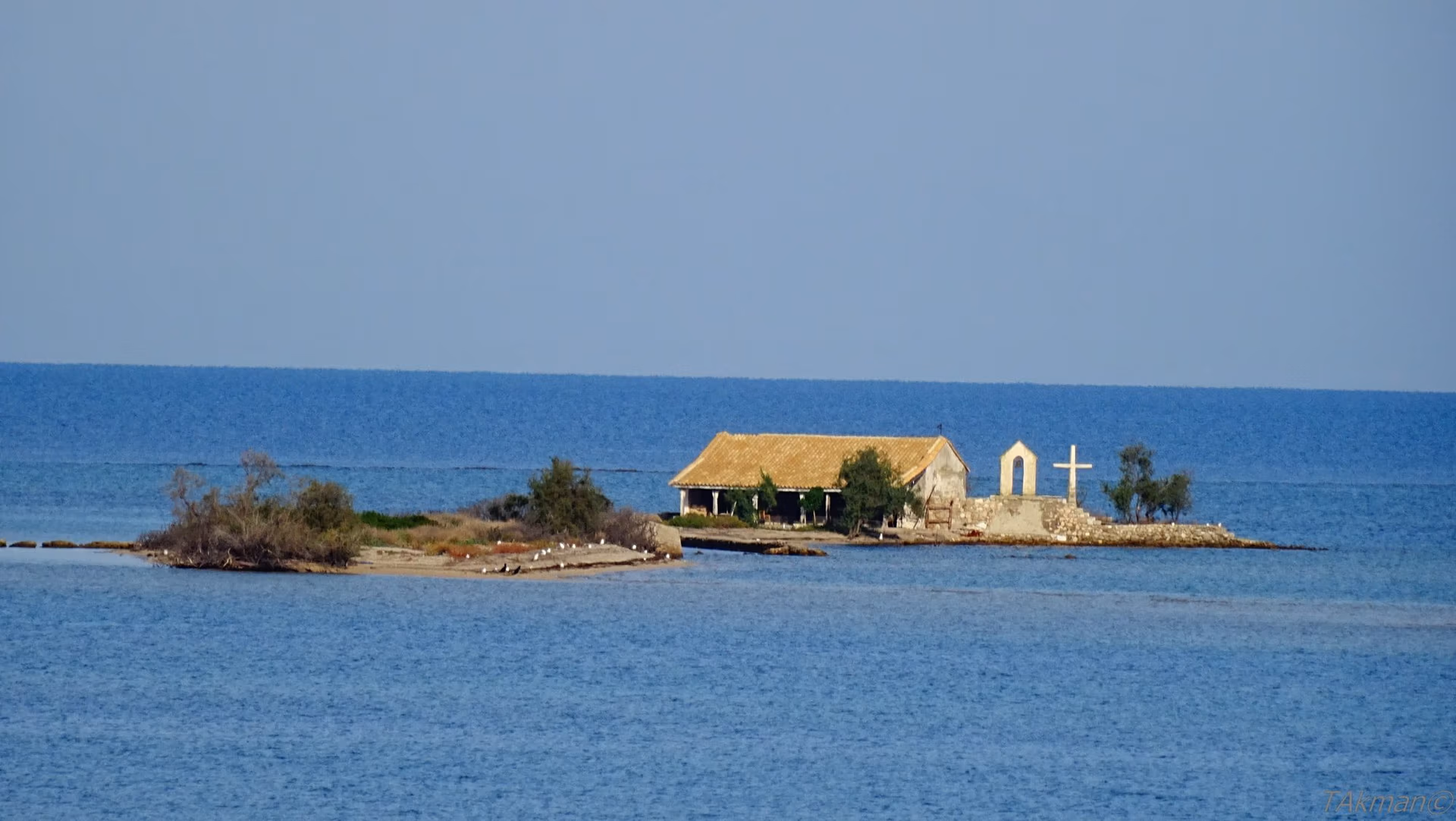 A low islet of sand and scrub in calm blue water, with a long single-storey chapel under an ochre tiled roof, a white arched bell gable and a white cross at its far end.