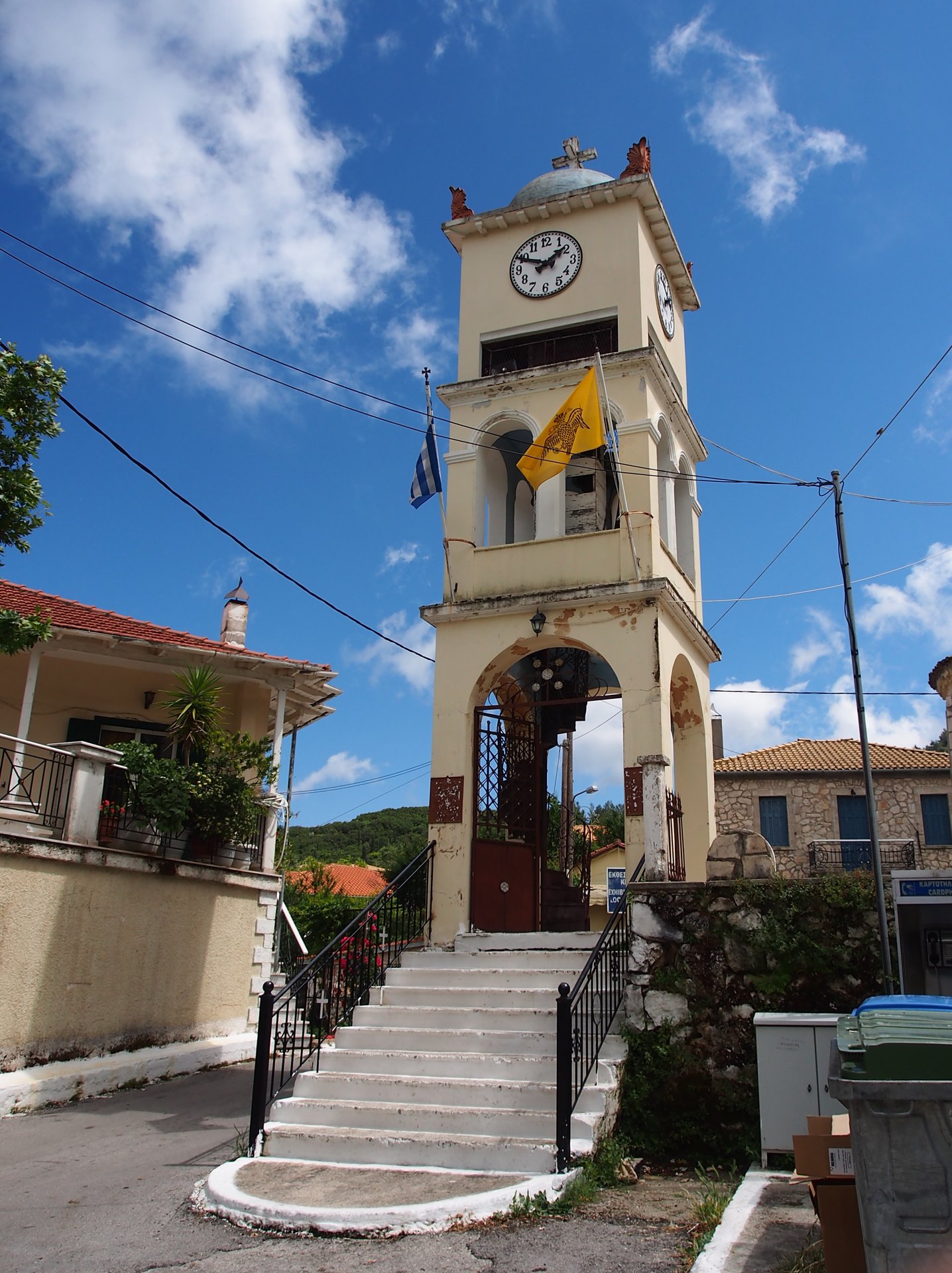 A pale stone clock tower with a Greek flag stands at the top of a flight of white-edged steps in Karya, under a blue sky.