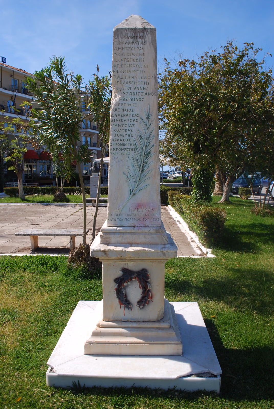 White marble obelisk memorial carved with names, on a stepped base in a lawn by the Lefkada town seafront