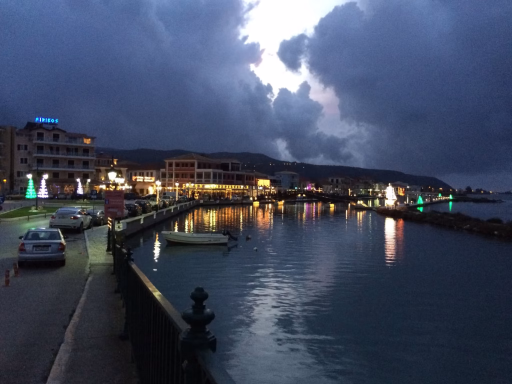 Lights of Lefkada Town reflected in the calm lagoon at dusk under heavy blue-grey clouds, with a small boat moored by the railing.
