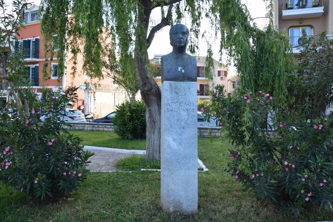 A dark bust of a bald man on a tall pale stone pillar inscribed with his name in Greek capitals, under a weeping willow in a small park with oleanders and town houses behind.