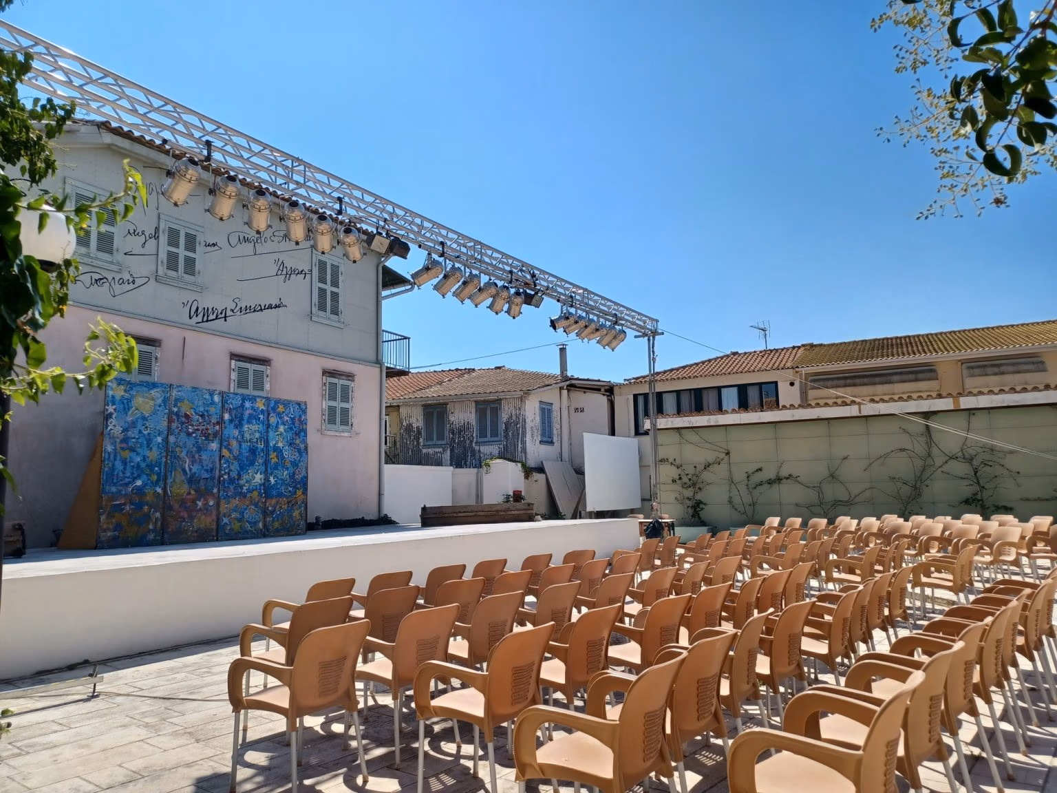 The courtyard garden theatre of the Angelos Sikelianos Museum, with rows of orange chairs facing a stage, beside a pale pink house whose gable carries painted handwritten signatures.