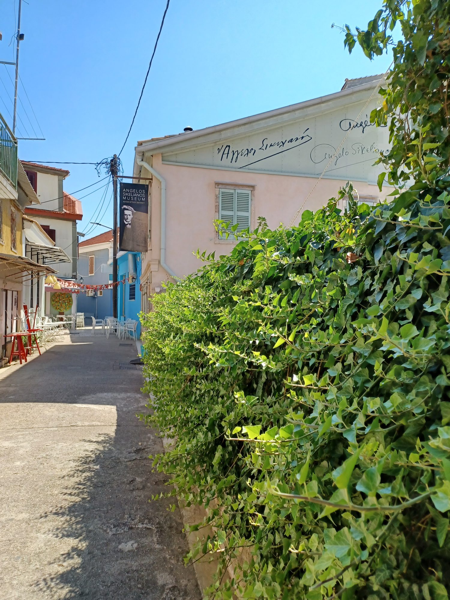 A narrow lane in Lefkada Town beside a pale pink two-storey house whose gable wall is painted with handwritten signatures, with a museum banner hanging at the corner.