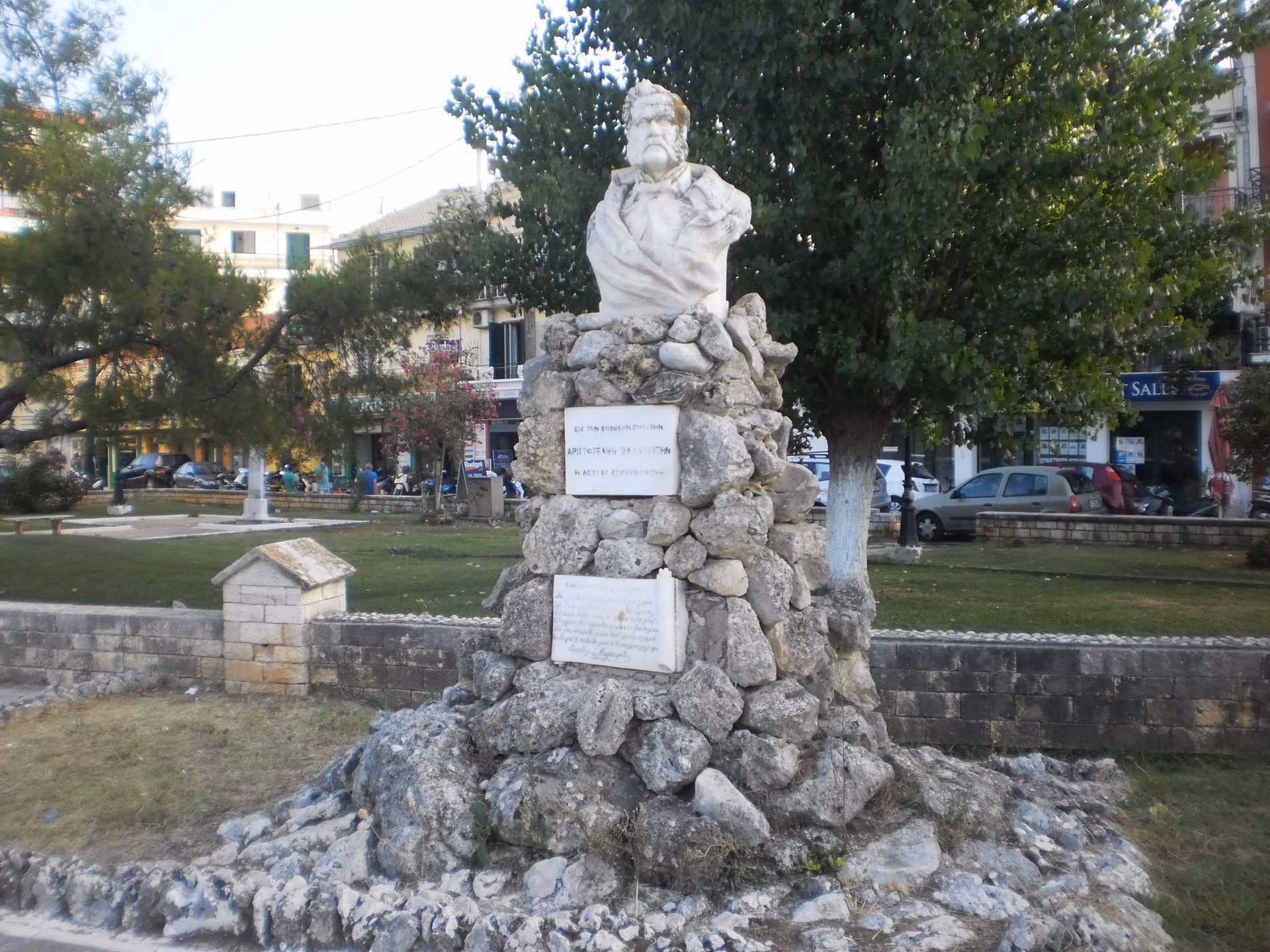 A white marble bust of a bearded man set on a cairn of rough stones with two inscribed plaques, in a small park with trees and town buildings behind.